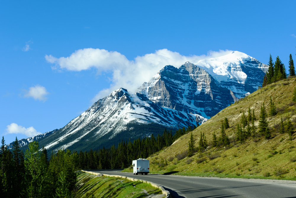 A white RV drives on a paved road through a mountainous landscape with snow-capped peaks and green trees under a clear blue sky.