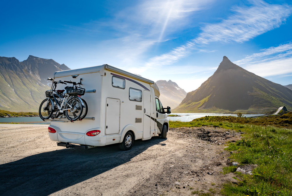 A white motorhome with bicycles mounted on the back is parked beside a scenic mountain lake under a clear blue sky.